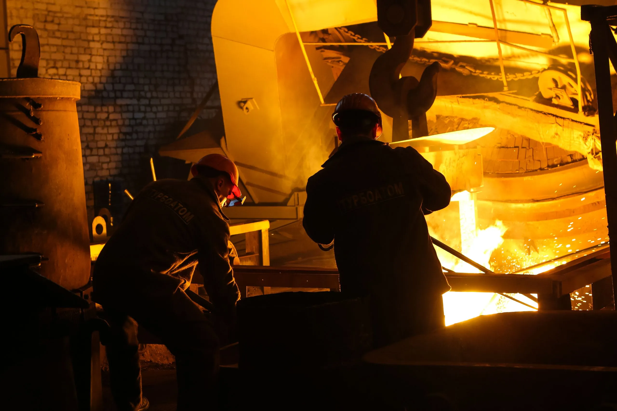 Two workers in hard hats at a furnace, molten metal glowing beyond them